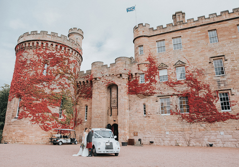 Bride and groom stand beside white wedding car in front of red leaf covered Dalhousie Castle